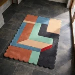 High angle view of the Soft Switch rug featuring a modern geometric color-block design with interlocking shapes in rust, light blue, and mint green, framed by a scalloped edge.