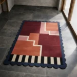 High angle view of the Stepped Field rug featuring a bold geometric design with burgundy, pink, and orange stepped blocks, accented by black and white stripes and a dark blue scalloped border.