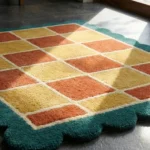 Close-up low angle view of the Sunbathe Quilt rug, showcasing the plush hand-tufted wool texture, the distinct square blocks in rust and mustard, and the intricately carved teal scalloped border.