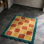 Perspective view of the Sunbathe Quilt rug placed on a dark tiled floor, highlighting the warm, sun-drenched checkerboard pattern and the contrasting teal scalloped edge.