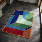 High angle view of the Sunroom Courtyard rug featuring a vibrant geometric color-block design with interlocking shapes in blue, green, red, and brown, framed by a rust scalloped border.