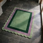 Perspective view of the Trimmed Forest rug placed on a dark tiled floor, showcasing the structured geometric border design and the organic light green scalloped edge illuminated by natural sunlight.