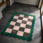 High angle view of the Verda Current rug featuring a stylish checkerboard pattern with alternating blush pink and charcoal grey squares, framed by a vibrant emerald green scalloped border.