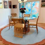 Perspective view of the circular Aurora Ring rug styled under a glass dining table with wooden chairs, showcasing its concentric circle design in rust, cream, and light blue against a hardwood floor.
