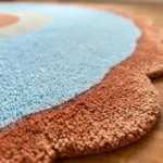 Close-up, low-angle view of the Aurora Ring rug's edge, highlighting the thick hand-tufted wool pile and the texture contrast between the light blue field and the wavy rust orange scalloped border.