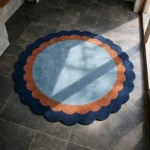 Perspective lifestyle view of the round Azure Halo rug placed on a dark tile floor, showcasing its distinct double scalloped border and contrasting blue and terracotta color palette in a sunlit entryway.