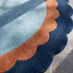 Close-up texture detail of the Azure Halo rug bathed in sunlight, highlighting the dense, plush hand-tufted wool pile and the clean, curved lines of the light blue, terracotta, and navy blue scalloped edges.