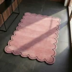 Perspective view of the blush pink scalloped rug styled in a sunlit modern room, showcasing the delicate wavy silhouette and stark black outline contrasting against a dark grey tile floor.