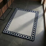 Perspective view of the Chain Frame square rug styled in a sunlit modern entryway, showcasing the elegant contrast of the navy scalloped border against dark tile flooring.
