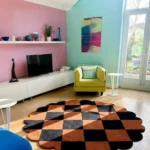 Perspective view of the circular Cinder Form rug on light hardwood floors, styled next to a teal velvet armchair and soft blankets, featuring a cream center with abstract charcoal organic shapes and a charcoal scalloped border.