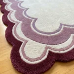 Close-up, angled view of the Elvare Bloom rug's corner, highlighting the thick, hand-tufted wool pile and the crisp carving that defines the nested scalloped borders in cream, pink, lavender, and plum.