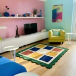 Perspective view of the Mosaic Verve square rug styled in a bright, eclectic living room with pastel pink and blue walls, resting on a light hardwood floor between a yellow armchair and a blue sofa.