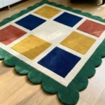 Angled close-up view of the Mosaic Verve rug on a hardwood floor, highlighting the dense hand-tufted wool pile, the crisp cream grid lines, and the plush, deeply carved green scalloped border.
