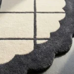 Close-up detail of the Orbital Grid rug bathed in sunlight, highlighting the dense hand-tufted wool pile, the crisp dark grey lines intersecting the cream base, and the plush sculpted edge.