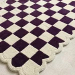 Perspective view of the Porcelain Rhythm checkerboard rug laid over a soft cream carpet, highlighting its distinct wavy scalloped edges and the rich contrast of the purple and cream geometric squares.