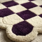 Close-up detail of the Porcelain Rhythm rug's corner, emphasizing the dense, hand-tufted wool cut pile and the seamless, deeply carved transition between the dark purple checkered squares and the plush cream scalloped border.