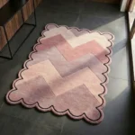 Perspective view of the Rose Chevron scalloped rug styled in a modern, sunlit entryway, showcasing its geometric pink and mauve zigzag design contrasting beautifully against a dark tile floor.