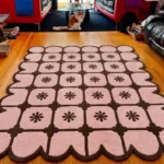 Perspective view of the pink and brown Roselle Lattice scalloped rug styled on a hardwood floor in a cozy living room, with a fluffy orange and white cat sitting in the background near a glass coffee table.