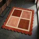Perspective view of the Sunset Panel square rug styled in a modern entryway, showcasing its warm rust and peach color-blocked geometric design contrasting against a dark grey tile floor in the sunlight.
