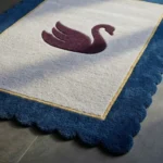 Angled close-up view of the Swan Crest rug bathed in sunlight, highlighting the dense, plush hand-tufted wool texture of the scalloped navy blue edge, the crisp gold border, and the central purple swan design.