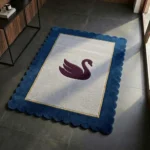 Perspective lifestyle view of the Swan Crest rug placed on a dark tile floor in a sunlit modern entryway, showcasing its elegant swan motif and distinct wavy blue border styled next to a sleek console table.