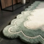 Close-up angled detail of the Sylvena Crest rug in natural sunlight, highlighting the dense hand-tufted wool pile, the crisply carved cream inner line, and the plush sage green scalloped edge.