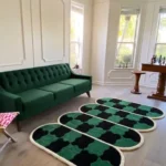 Perspective view of the Verdure Drift wavy checkerboard rug styled on a light hardwood floor in a sunlit, elegant living room, beautifully complementing a classic tufted emerald green velvet sofa.