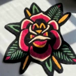 Flat top-down view of the American Traditional Rose Rug, clearly displaying the entire repeating traditional American tattoo flash pattern of two red roses, blue accents, green leaves, and black dotted stars on a cream center, framed by a distinctive dark scalloped border with red trim.