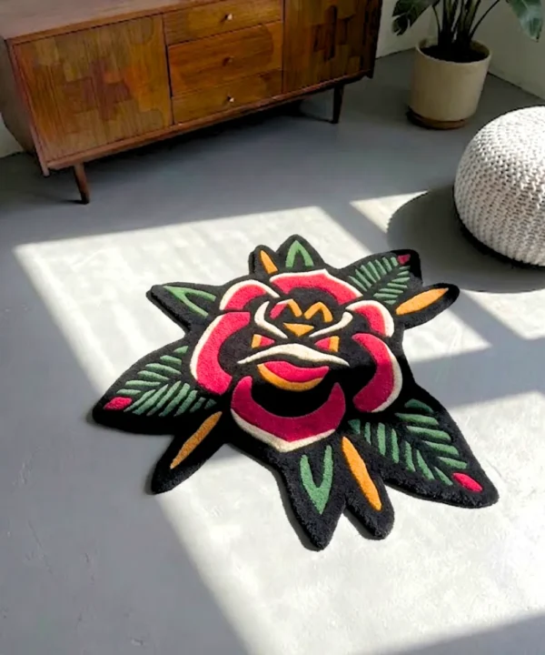 Perspective view of the rectangular American Traditional Rose Rug, featuring a traditional tattoo flash-style repeating pattern of two red roses and stars on a cream background with a scalloped black border, styled on a modern concrete floor next to a textured white pouf and a mid-century wooden cabinet bathed in natural sunlight.