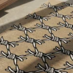 Angled view of the tan Barbed Wire tattoo flash rug resting on a smooth concrete floor, beautifully bathed in natural sunlight with sharp architectural shadows cast across its repeating pattern.