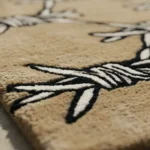 Close-up macro detail of the Barbed Wire rug's edge in the sunlight, highlighting the dense hand-tufted tan wool pile and the crisp carving of the black and white design.