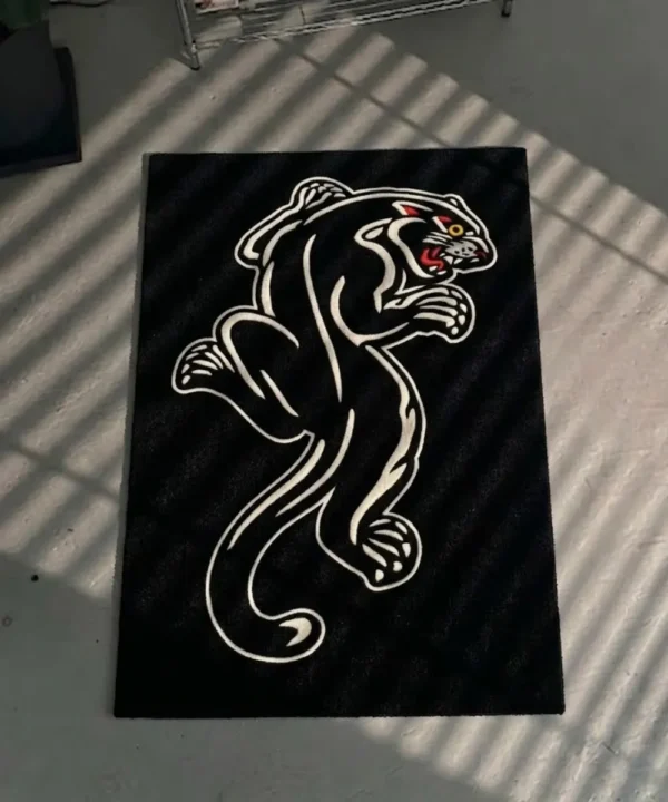 Top-down flat view of the die-cut Black Panther Tattoo Rug, resting on a grey concrete floor, showcasing its irregular shape, crouching panther silhouette, and vibrant red, yellow, and blue traditional tattoo-style face and border accents.