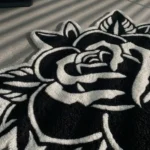 Angled close-up view of the Black Veil black and white rose rug placed on a smooth grey concrete floor, beautifully illuminated by dramatic slatted window blind shadows.