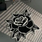 Top-down view of the Black Veil rose-shaped rug resting on a concrete floor with striking window blind shadows cast across its surface, emphasizing the bold contrast of the white carved lines on the black wool background.
