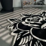 Perspective view of the Black Veil tattoo flash rose rug styled in a modern studio space on a grey concrete floor, positioned near a potted plant and a metal shelving unit displaying pop culture collectibles.