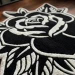 Close-up angled view of the Black Veil rose rug resting on a dark hardwood floor, highlighting the thick hand-tufted wool pile and the crisp 3D carving of the white floral outlines against the solid black base.