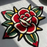 Top-down close-up view of the Classic Rose Tattoo Rug, emphasizing the bold traditional American tattoo flash style, red petals, and green leaf details in natural light.