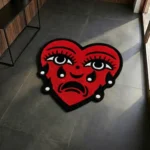 Top-down flat view of the die-cut Crying Heart rug, featuring a bold traditional tattoo flash design of a red heart with a face, striking blue tears, and a solid black border.