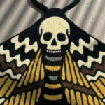 A perspective shot showing the die-cut Death Head Moth traditional tattoo rug placed on a grey concrete floor, positioned in front of a warm wooden cabinet.