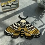 Top-down view of the hand-tufted Death Head Moth rug, in traditional tattoo style with gold, black, and cream colors, lying flat on a grey concrete floor with natural sunlight shadows.