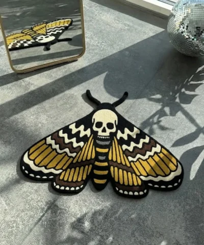 Top-down view of the hand-tufted Death Head Moth rug, in traditional tattoo style with gold, black, and cream colors, lying flat on a grey concrete floor with natural sunlight shadows.