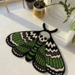 Angled perspective view of the die-cut Death Moth traditional tattoo rug styled on a bright white floor next to a green potted plant, showcasing its intricate green wings and stark white skull design.