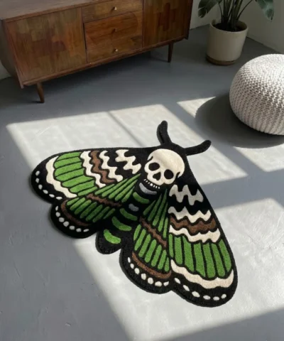 High-angle view of the Death Moth neo-traditional tattoo flash rug resting on a smooth grey concrete floor next to a white pouf, with dramatic window shadows cast across its skull and wing details.