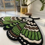 Low-angle perspective view of the Death Moth rug bathed in natural sunlight on a white floor, emphasizing the thick, plush texture of the hand-tufted green, brown, and white patterned wings.