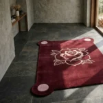 Wide perspective view of the Gothic Rose Emblem rug bathed in sharp sunlight and shadows, placed on a dark stone tile floor in a modern room with a wooden bench and large windows.