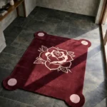 Perspective view of the rectangular Gothic Rose Emblem rug featuring a deep burgundy background, a central cream traditional tattoo rose, and four light pink circular corner accents, styled on a dark slate floor in bright natural sunlight.