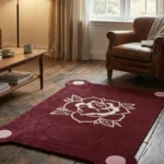 Perspective view of the burgundy Gothic Rose Emblem rug styled on a rustic hardwood floor in a cozy living room, positioned near a classic brown leather armchair and a wooden coffee table.