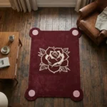 High-angle top-down view of the Gothic Rose Emblem rug resting on dark rustic wood planks, perfectly showcasing its rectangular shape with rounded pink corner extensions and central cream rose motif, framed by cozy den furniture.