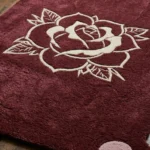 Close-up macro detail of the Gothic Rose Emblem rug, highlighting the dense hand-tufted burgundy wool pile, one of the pink circular corner extensions, and the crisp 3D carving of the central cream rose design.
