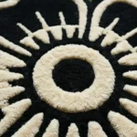 Close-up corner detail of the hand-tufted wool Inked Garden Rug, highlighting the dense texture of the natural fiber pile, the detailed repeating traditional tattoo floral pattern, and the precise stitching of the dark scalloped border on a textured concrete surface.